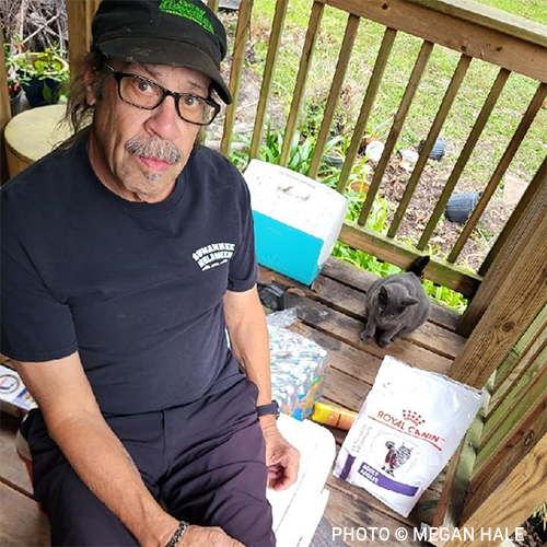 Image shows Suwannee County resident on the porch of his damaged home with Silver, his six-year-old cat, next to a bag of donated Royal Canin cat food.